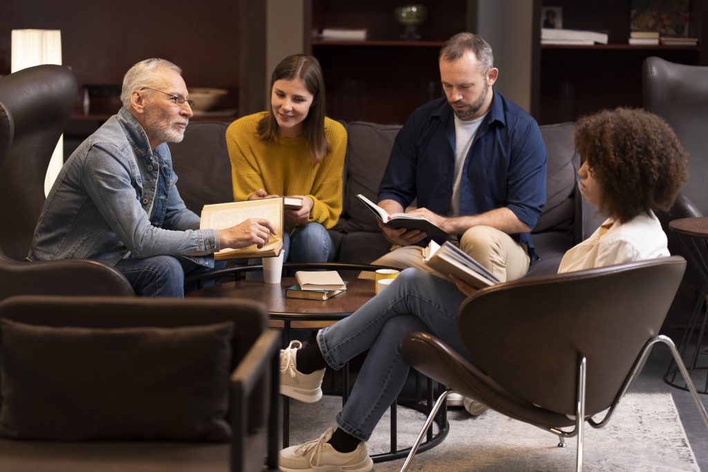 Four people sit in a lounge area, engaged in discussion and reading books around a coffee table with notebooks and coffee cups, sharing ideas on how to minimize family conflicts after your passing.