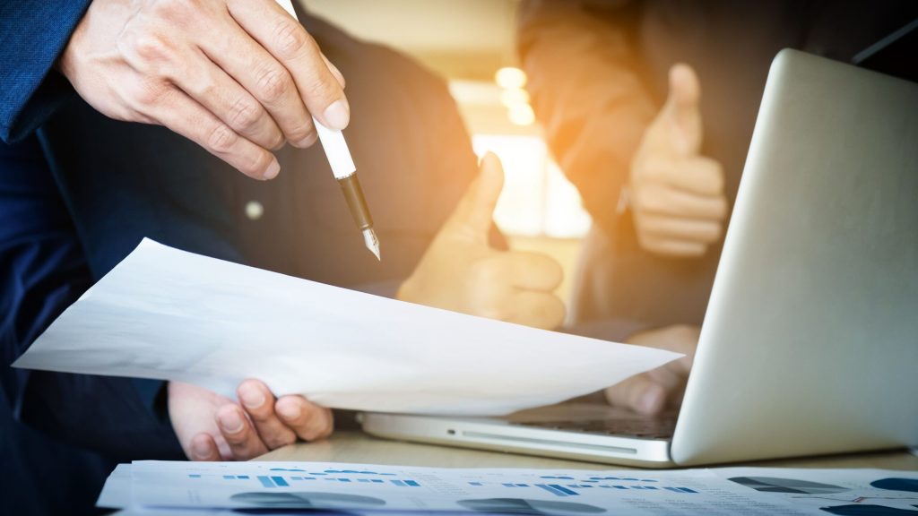 Two people at a desk review a document with charts—perhaps comparing Will Vs Trust Which One Do You Really Need—one holds a pen and paper, while the other gives a thumbs-up near an open laptop.