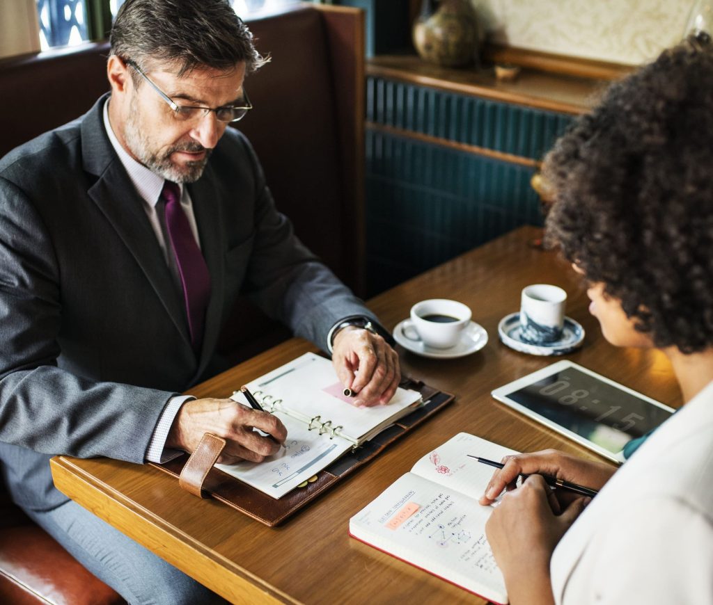 Two people in business attire sit at a table with notebooks, discussing planificación de la incapacidad. A tablet, papers, and cups of coffee are also on the table as they review important details.