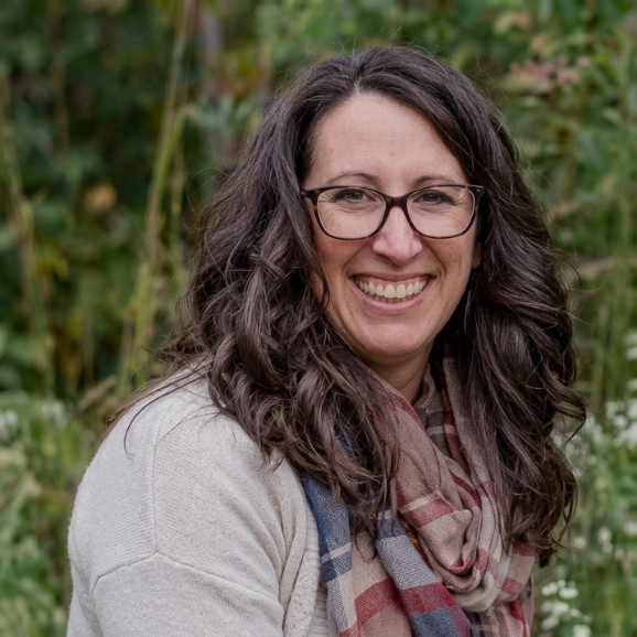 Kathryn Putnam, a woman with long brown hair and glasses, wears a beige sweater and plaid scarf, smiling outdoors with greenery in the background.