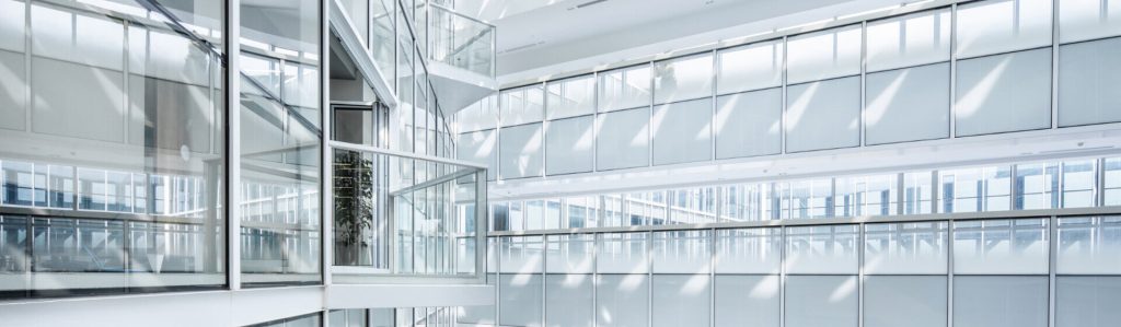 Interior view of a modern building with large glass windows, white walls, and sunlight casting diagonal shadows across surfaces.