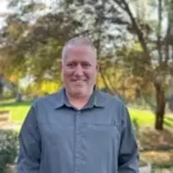 A man with short hair wearing a gray button-up shirt stands outdoors in a park with trees and greenery in the background.