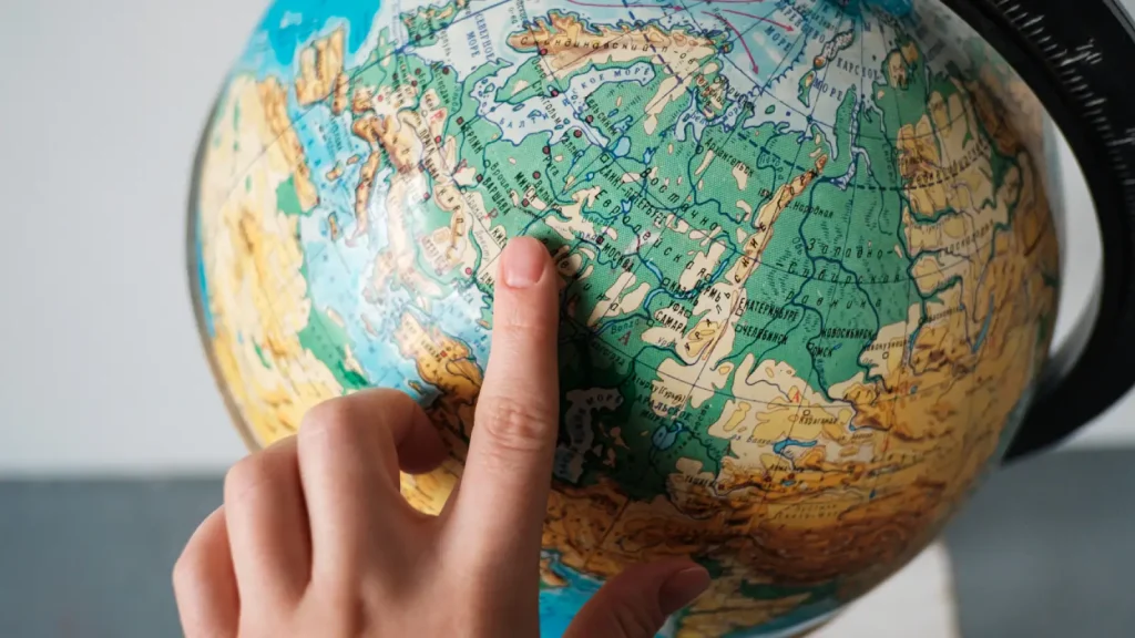 A hand points to a location on a globe, focusing on an area in northern Asia, with visible countries and geographical details.