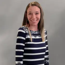 A woman with straight brown hair, wearing a navy and white striped long-sleeve top, stands in front of a plain gray background.