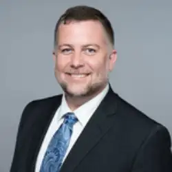 A man in a suit and tie poses for a professional headshot against a plain gray background.