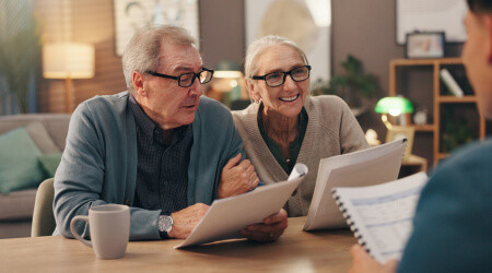An older couple sits at a table holding documents and talking to a person across from them, who is also holding paperwork.