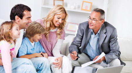 A man in a suit discusses papers with a smiling family of four, including two children, seated together in a bright living room.